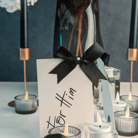 Name card with black bow and 'For Him' text on a table with candles and flowers.