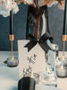 Name card with black bow and 'For Him' text on a table with candles and flowers.