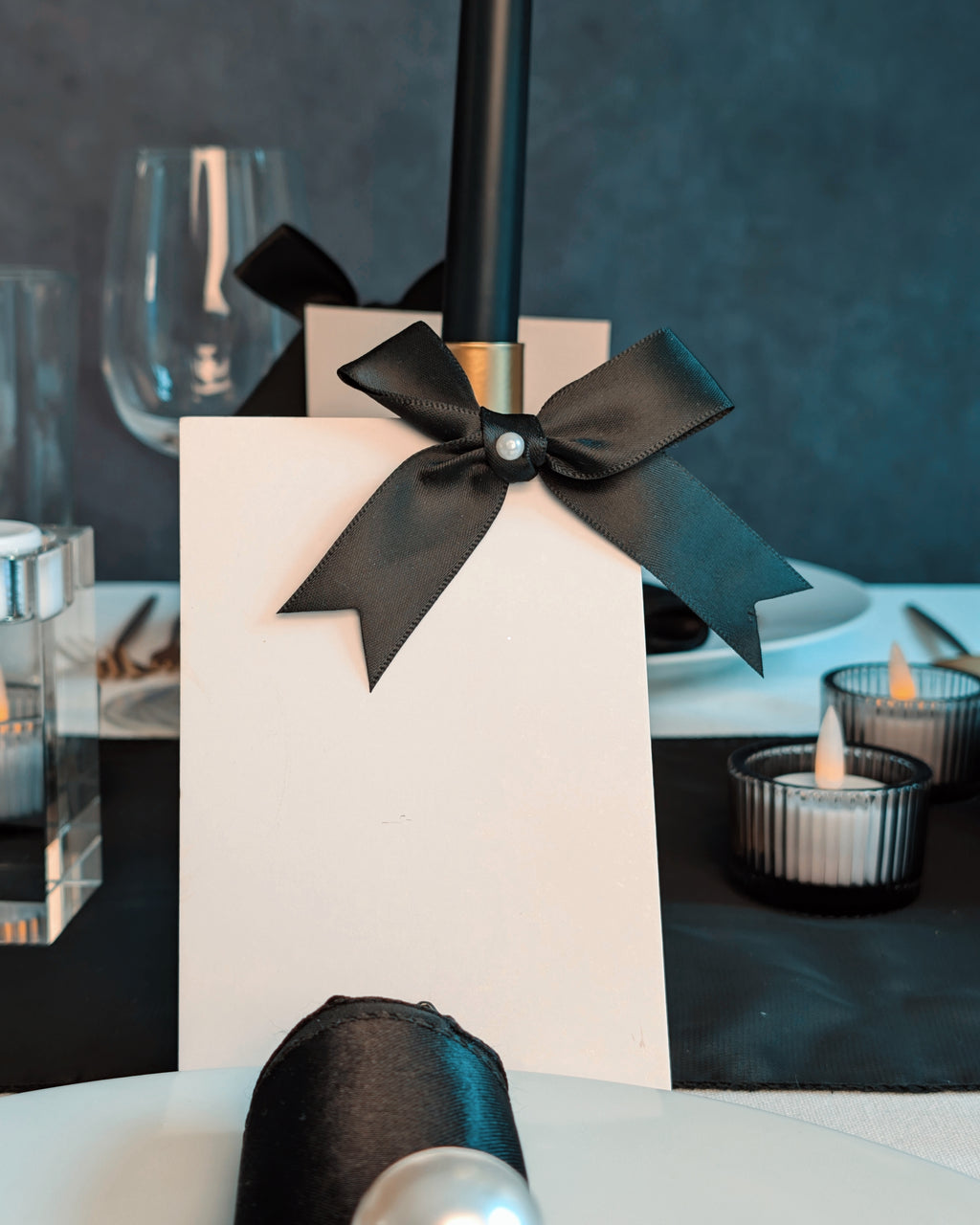 White name card with a black ribbon on a table setting with candles and glasses. For a black and white tablescape