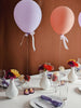 Elegant table setting with purple and peach balloons tied to colorful flower vases. Soft candlelight and folded napkins create a festive ambiance.