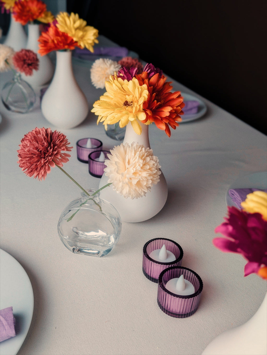 White vases with vibrant flowers in red, yellow, and orange are arranged on a table with purple candle holders, creating an elegant and cheerful atmosphere.