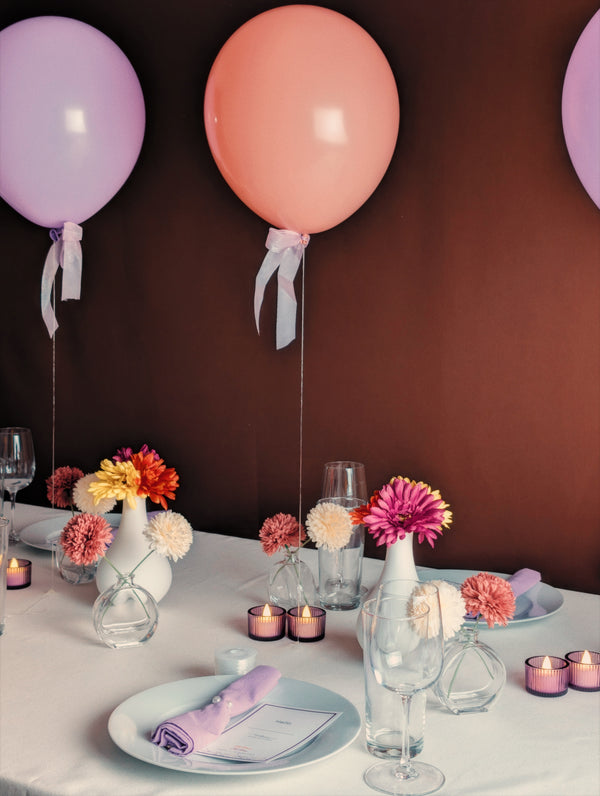 A table setting with pink and purple balloons against a brown wall, adorned with colorful flowers in vases, candles, wine glasses, and lilac napkins.