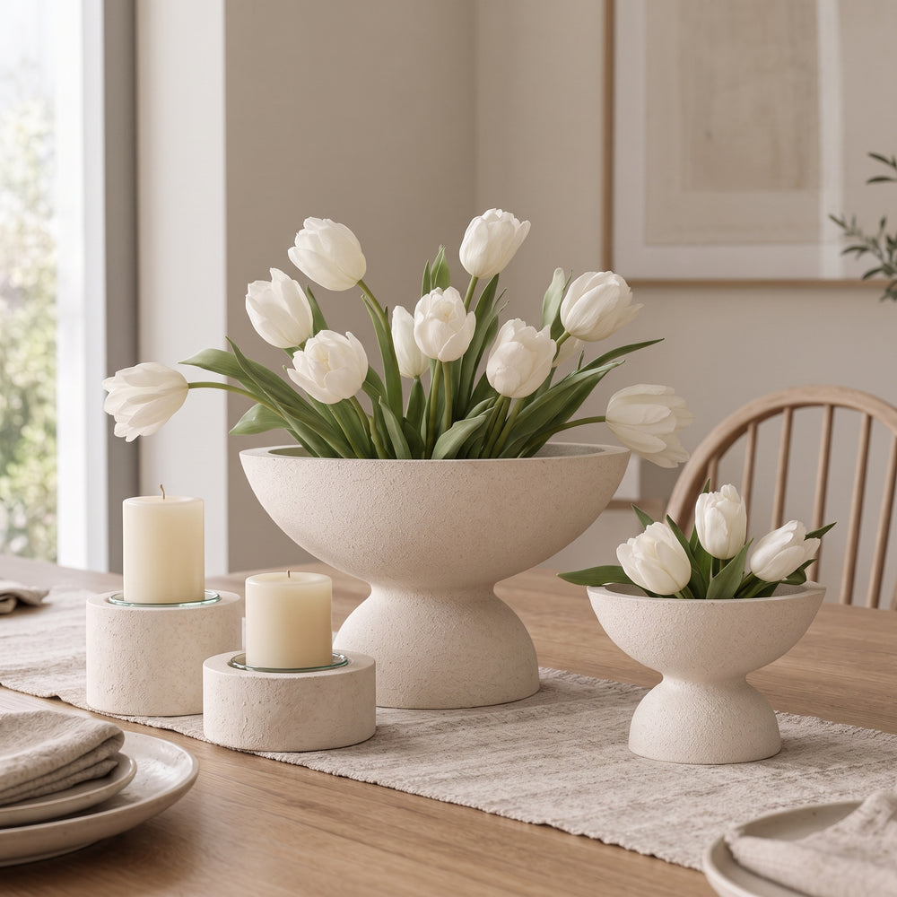A styled dining table scene with three matte cream ceramic pedestal flower bowls holding white tulips, alongside two matching cream pillar candles on small round holders. The setting includes a wood chair, framed wall art, and a window in a softly lit interior.
