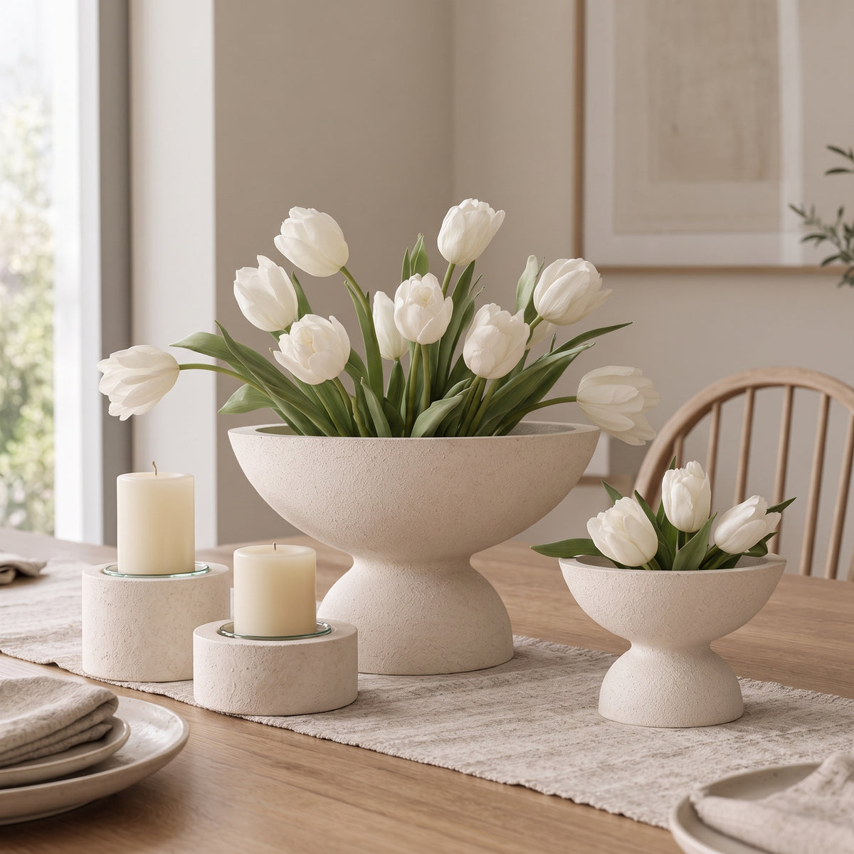 A styled dining table scene with three matte cream ceramic pedestal flower bowls holding white tulips, alongside two matching cream pillar candles on small round holders. The setting includes a wood chair, framed wall art, and a window in a softly lit interior.