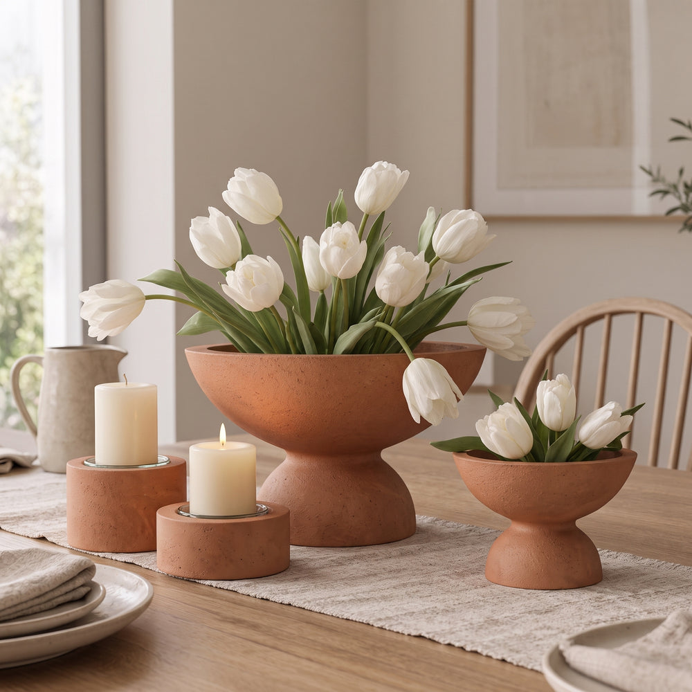 Terracotta-colored ceramic tulip holders on a dining table, with a large pedestal bowl filled with white tulips in the center and two smaller matching holders on either side. The scene is styled in a warm, neutral-toned dining room with candles, plates, and a wooden chair.