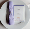 A white plate with a neatly folded lavender napkin adorned with pearl-like buttons, next to a welcome card with the text "Hello" and floral accents.