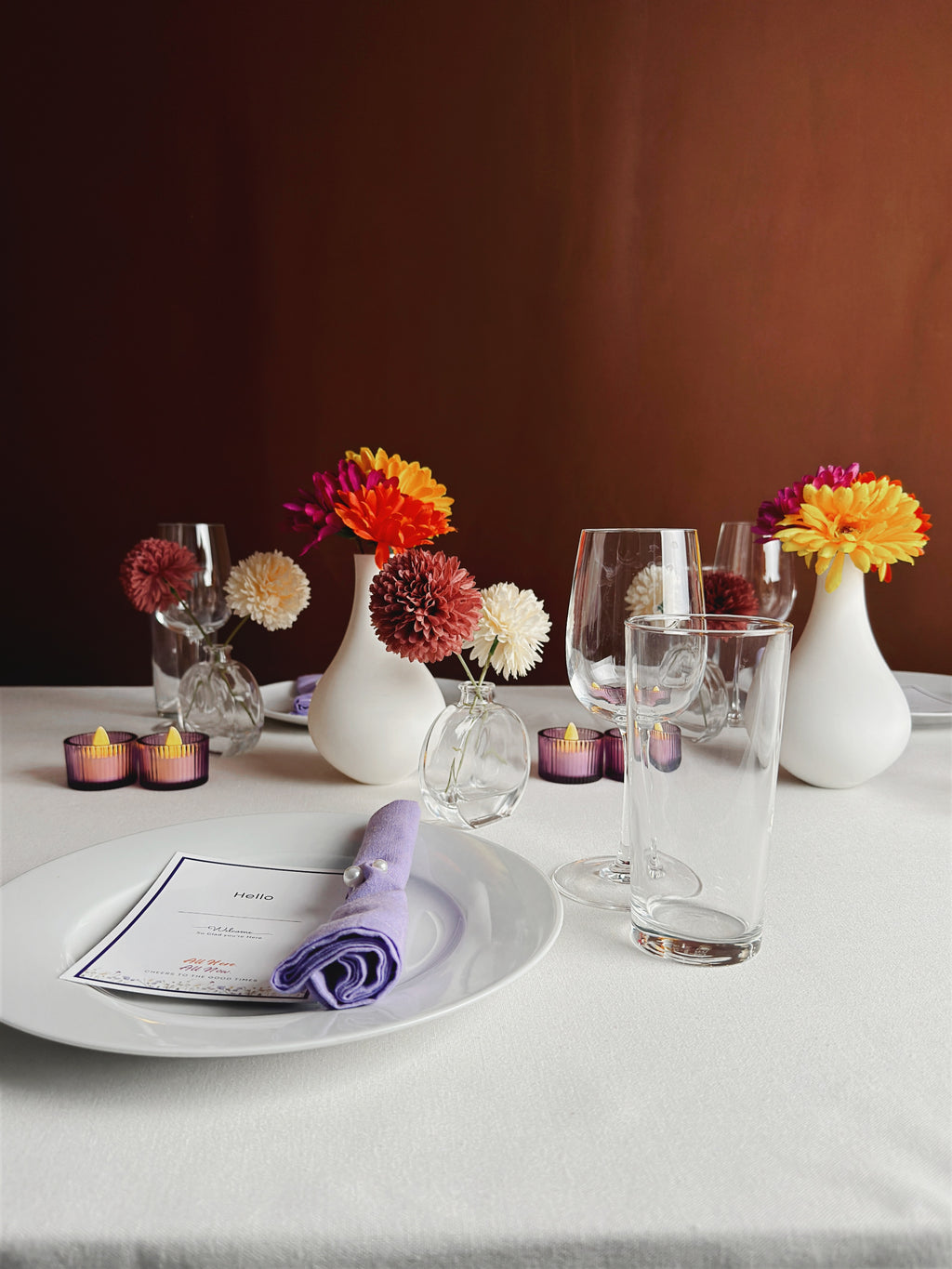 Elegant dining table setting with white dishes, purple napkins, and glassware. Colorful flowers in white vases and warm candles create a cozy atmosphere.