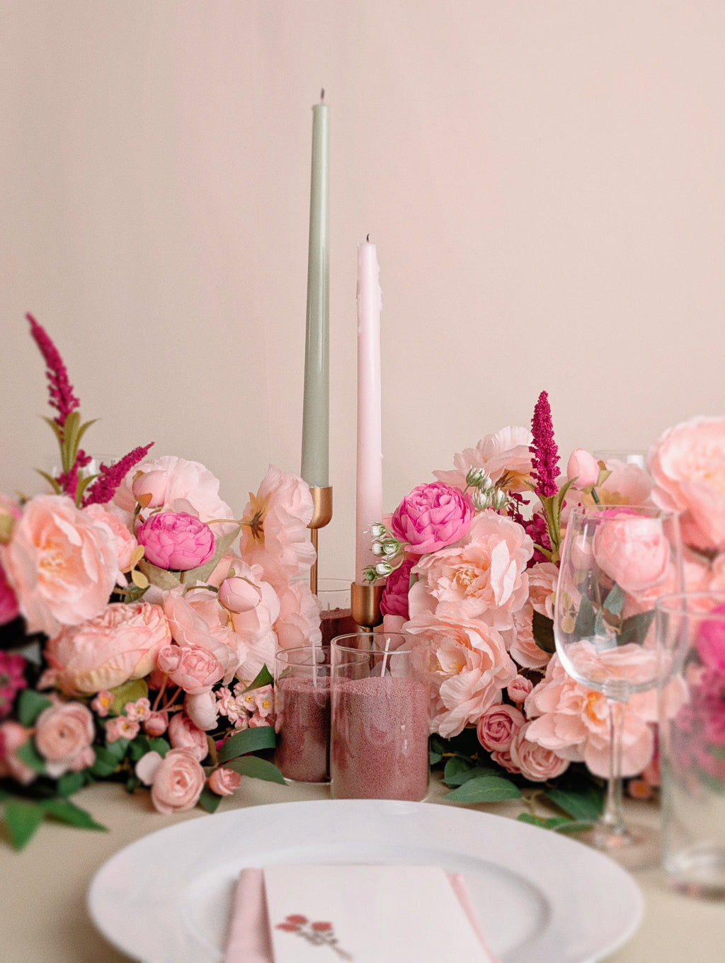 Table setting with pink flowers, candles, and glasses on a beige background